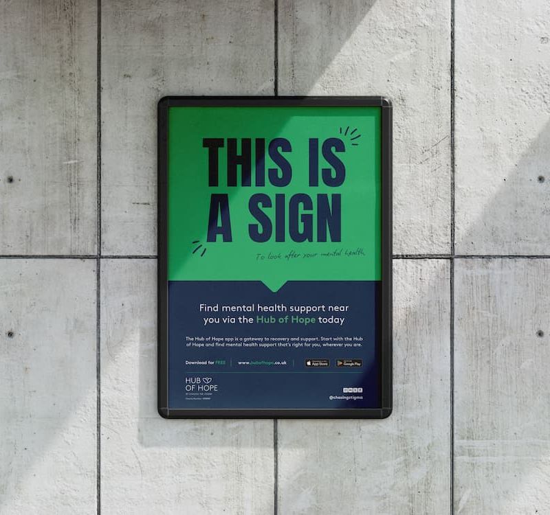 A rectangular, green-and-black poster mounted on a concrete wall. The top half reads “THIS IS A SIGN” in bold letters, with a smaller subtitle: “to look after your mental health.” The lower portion encourages finding mental health support through the Hub of Hope, showing a URL and app store icons